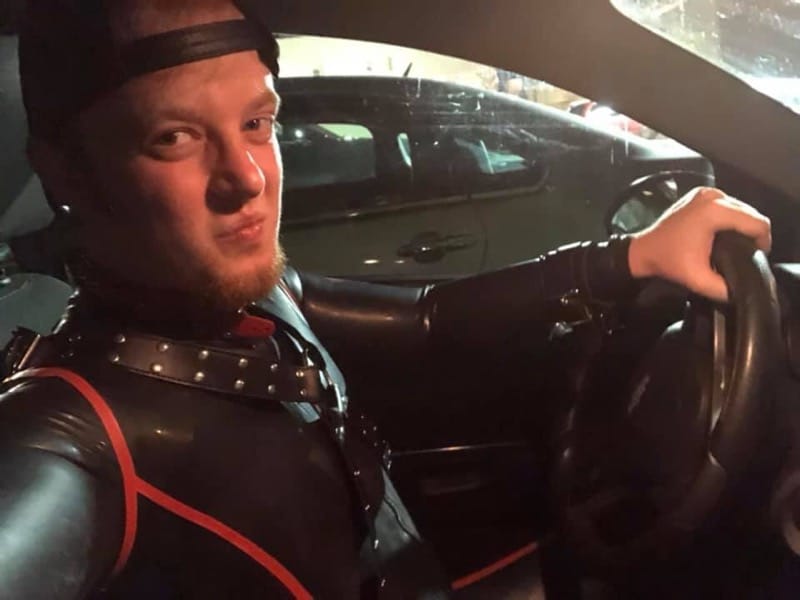 photograph of luke wenke driving a car in a latex bondage suit and a backwards baseball cap